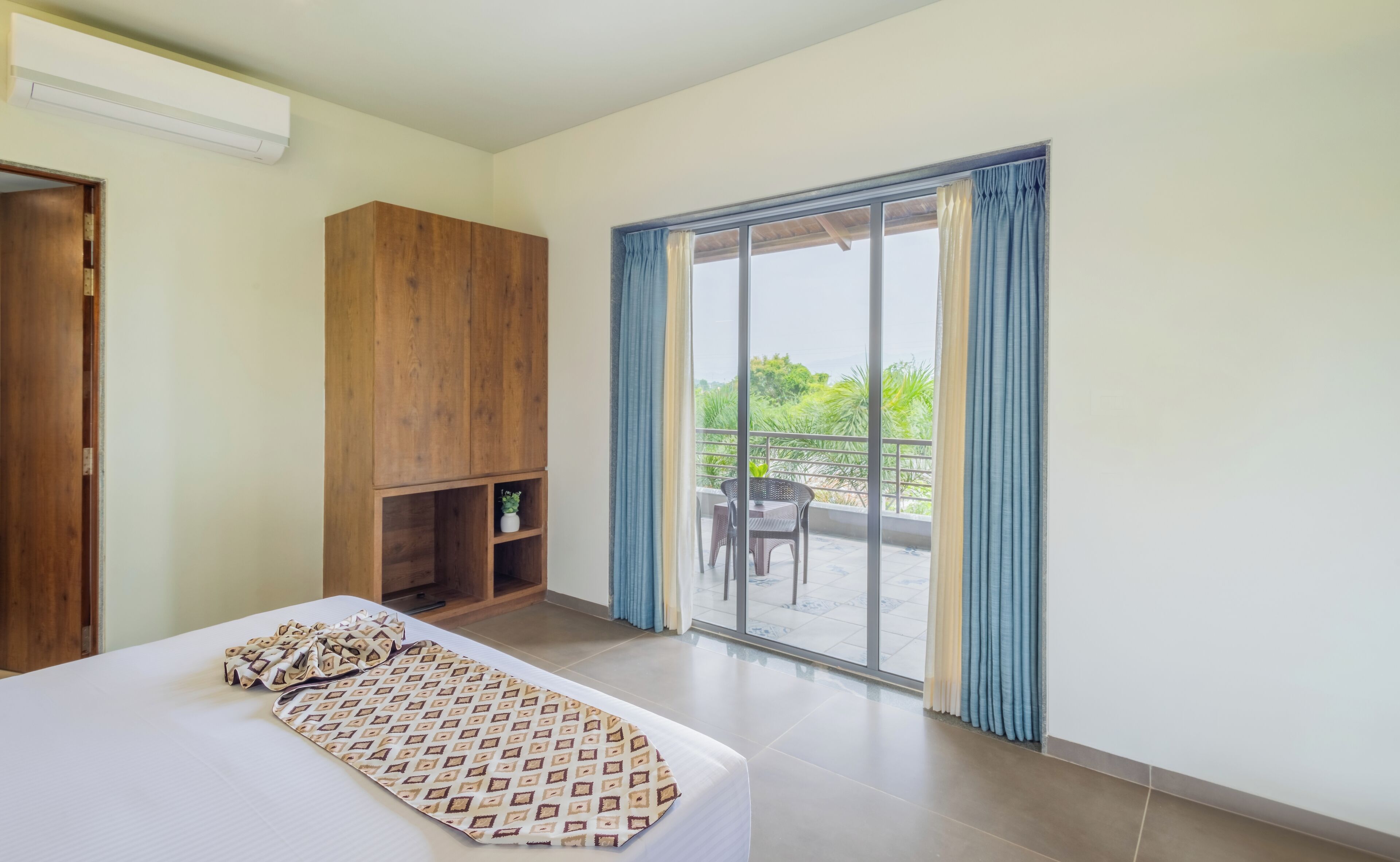 Een hotelkamer met een bed, een houten kledingkast, een klein plankje met een plant en een balkon met een tafel en stoelen.