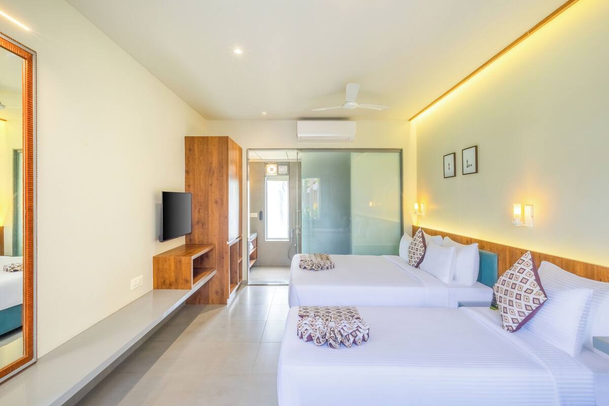 Quarto de hotel moderno com duas camas, televisão, espelho e corredor que dá para um banheiro.