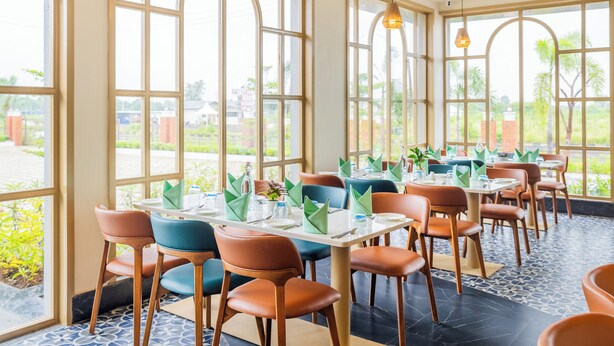 Un restaurante con amplios ventanales, varias mesas dispuestas con servilletas verdes y un suelo de baldosas.