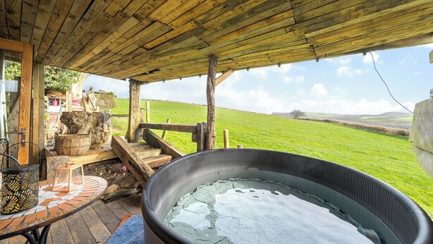 Une terrasse en bois avec un jacuzzi, une table dont le plateau est recouvert de carreaux, et une vue sur un champ verdoyant.