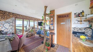 Un intérieur chaleureux avec un mur en pierre, un tronc d’arbre servant de pièce maîtresse, un canapé, un coin repas et une cuisine dotée d’étagères.