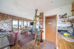 A cozy interior with a stone wall, a wooden tree trunk centerpiece, a sofa, a dining area, and a kitchen with shelves.