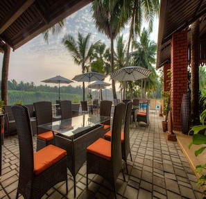 An outdoor patio with wicker furniture and umbrellas, overlooking a lush landscape with palm trees. - RiverRoost Resorts (Mangaluru)