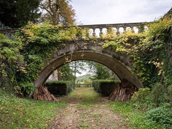 An arched stone bridge with ivy overgrown on the sides, surrounded by greenery and a path leading through it.