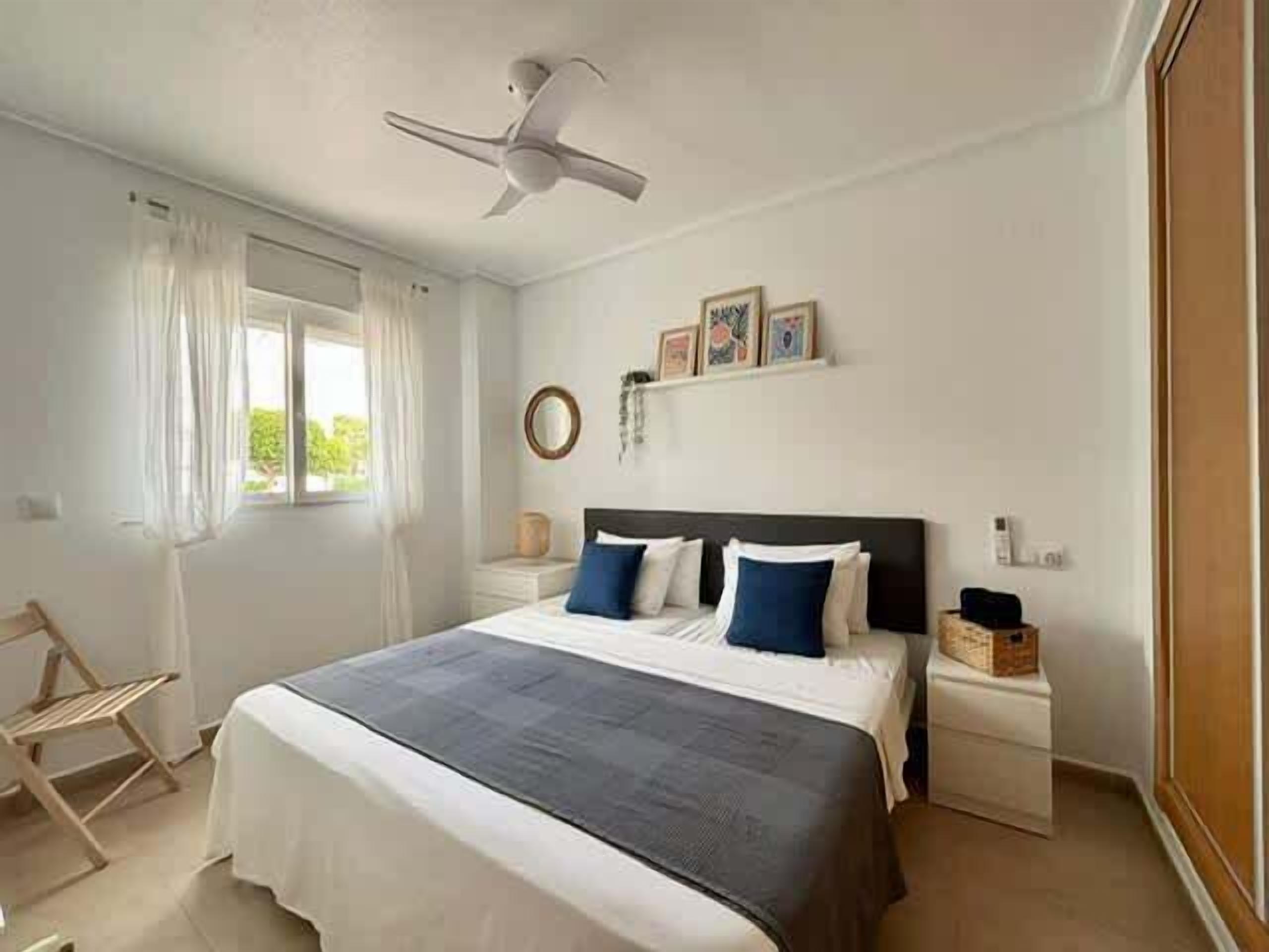 Une chambre à coucher comprenant un lit, un ventilateur de plafond, une fenêtre avec des rideaux, une table de chevet et une commode.