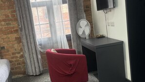 A room with a brick wall, a window with curtains, a red chair, a desk with a fan, and a TV mounted on the wall. - Studio - Sleeps 2 - Parking - Wifi (Nottingham)
