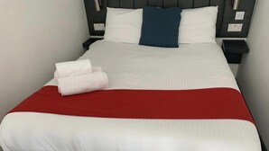 Un lit bien fait, avec une literie blanche et rouge, un oreiller bleu et deux serviettes blanches pliées.