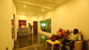 A hotel lobby with a reception desk labeled "Lé Apex," a seating area with a sofa and a coffee table, and a large window with a view. - Le Apex (Vanur)