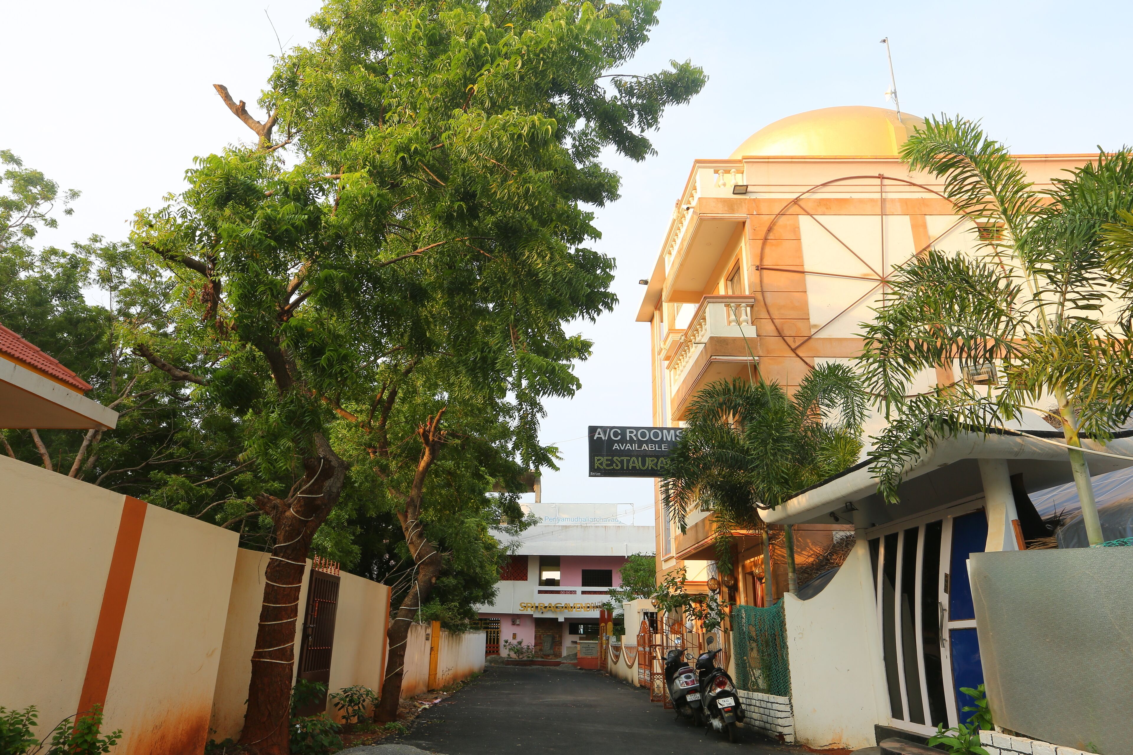 A narrow street with a building featuring a large golden dome, a sign for A/C rooms and a restaurant, and a parked scooter.
