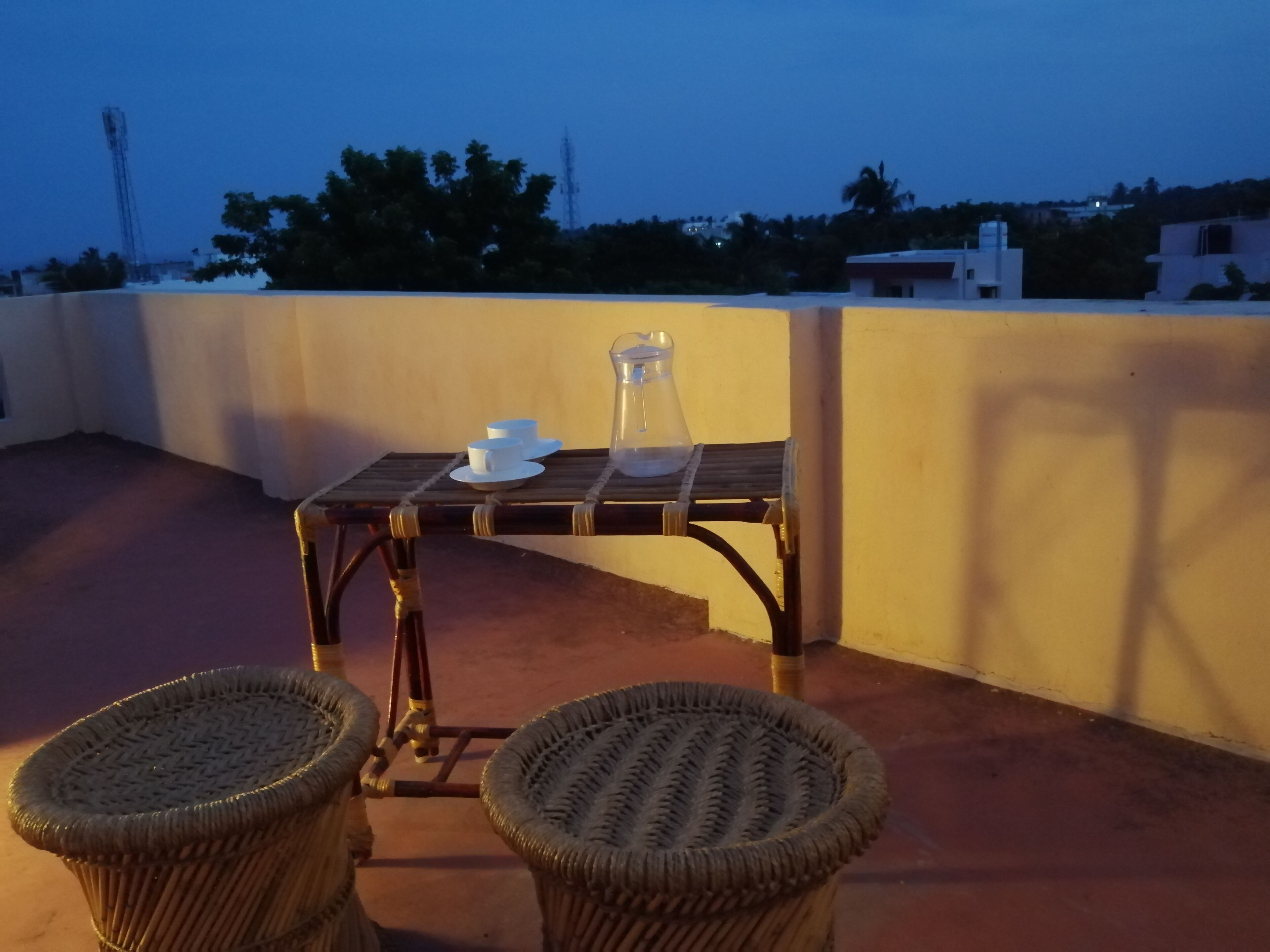 A rooftop terrace with a table set for two, two wicker chairs, and a view of the cityscape at dusk.