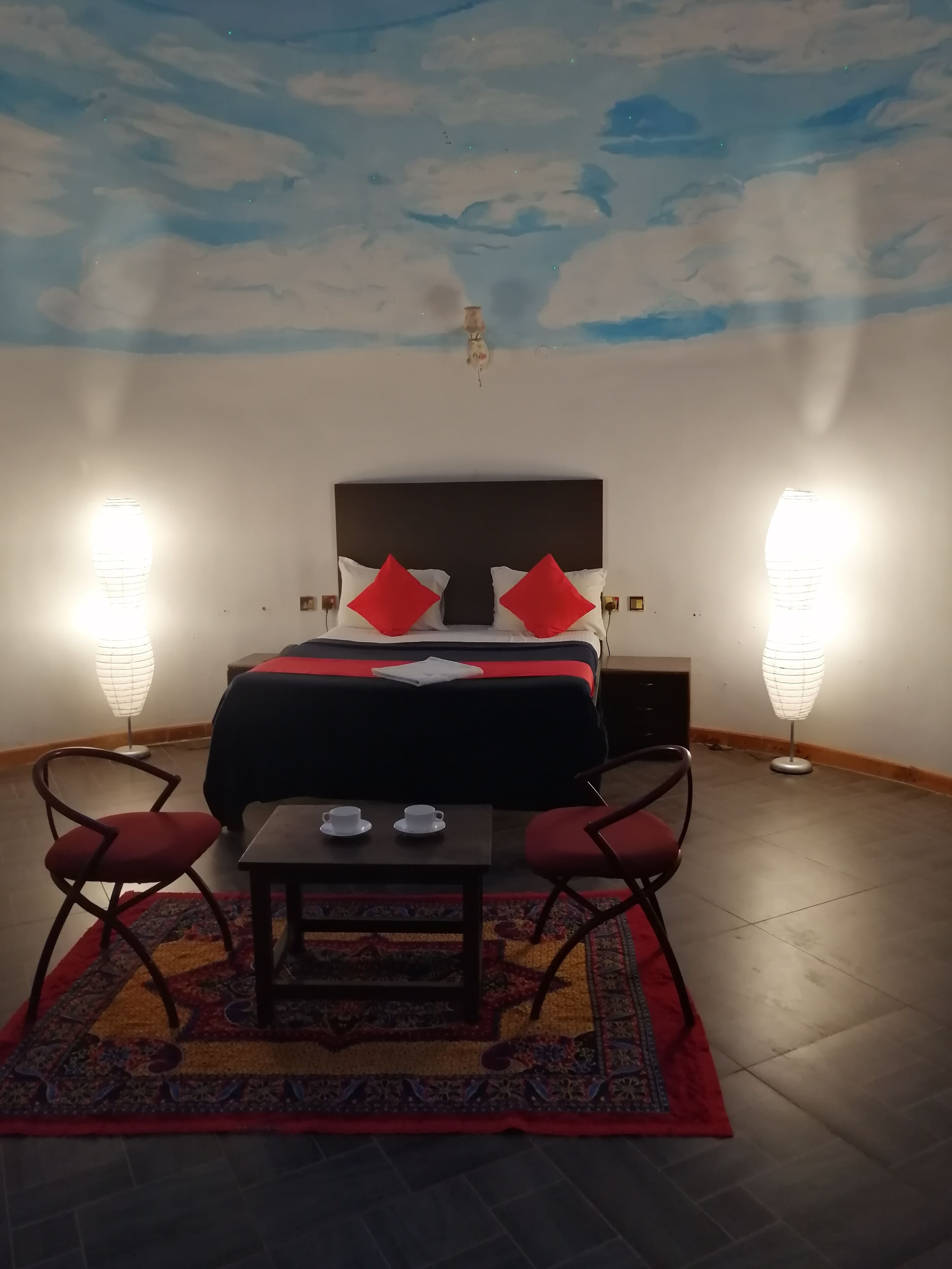 A hotel room with a bed, two red chairs, a small table with cups, and two floor lamps.