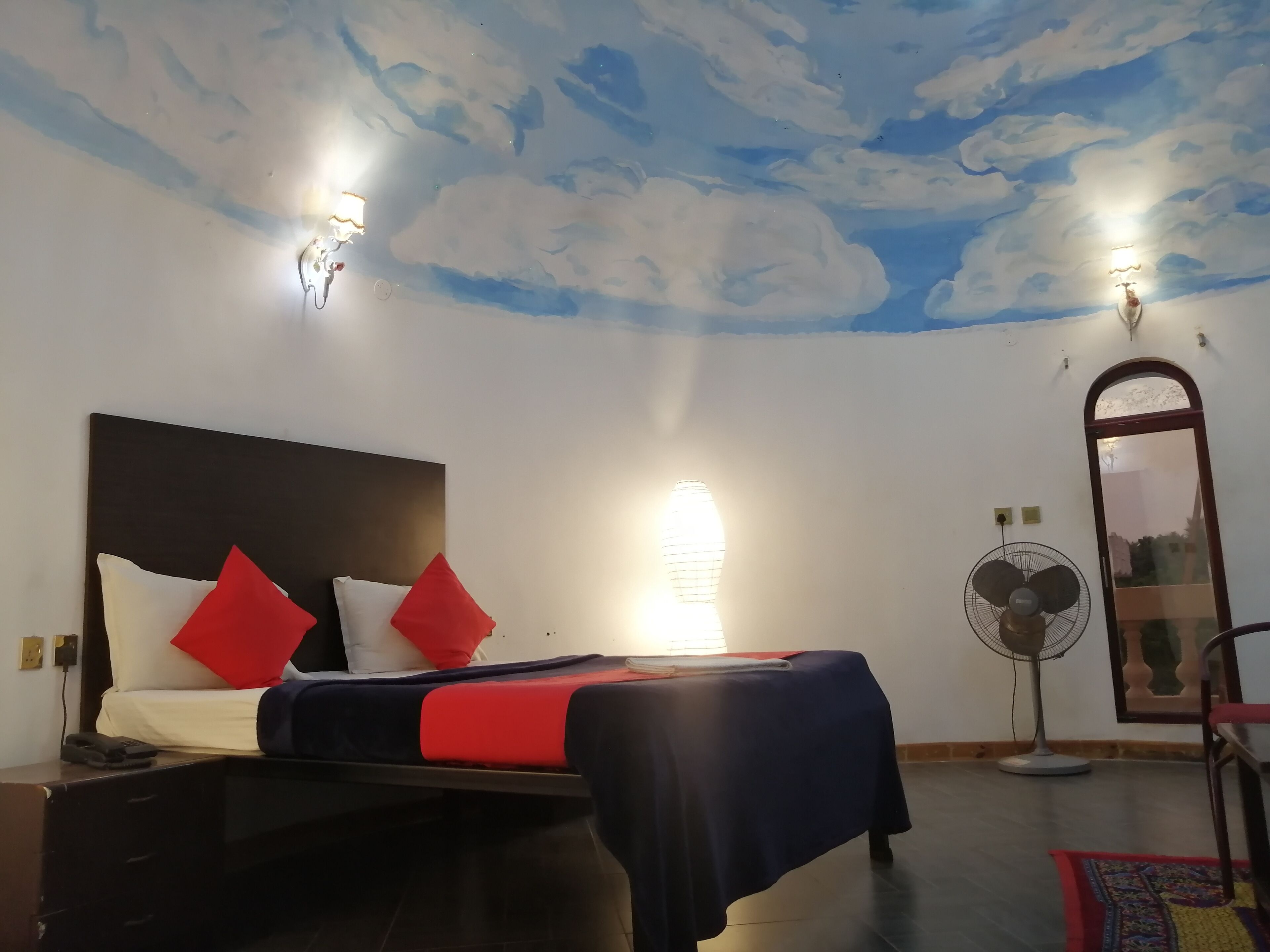 Une chambre d’hôtel avec un lit, un ventilateur sur pied et un miroir au mur.