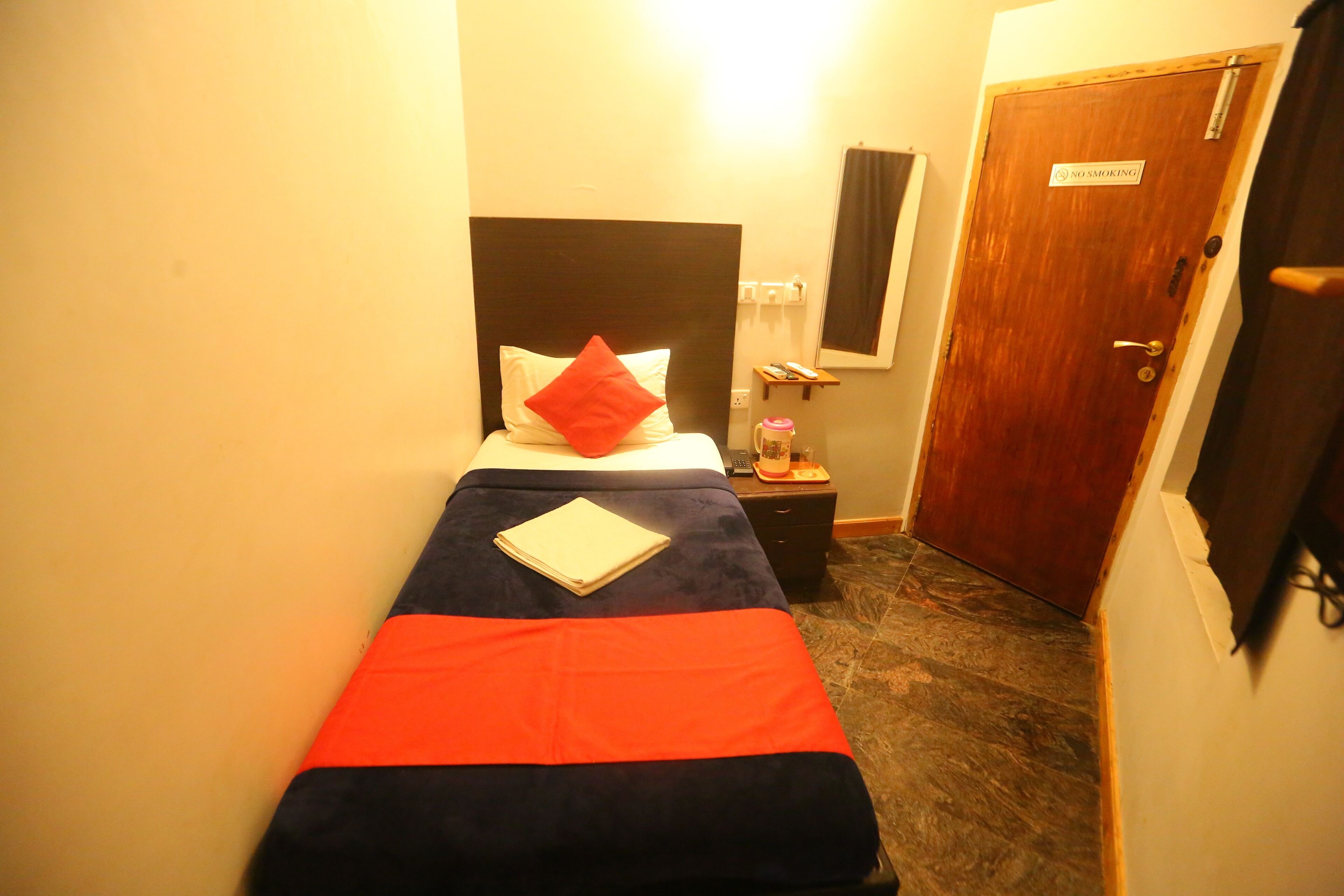 Habitación de hotel estrecha con una cama individual, una mesita de noche, una pequeña mesa con lámpara y una puerta con letrero de prohibido fumar.