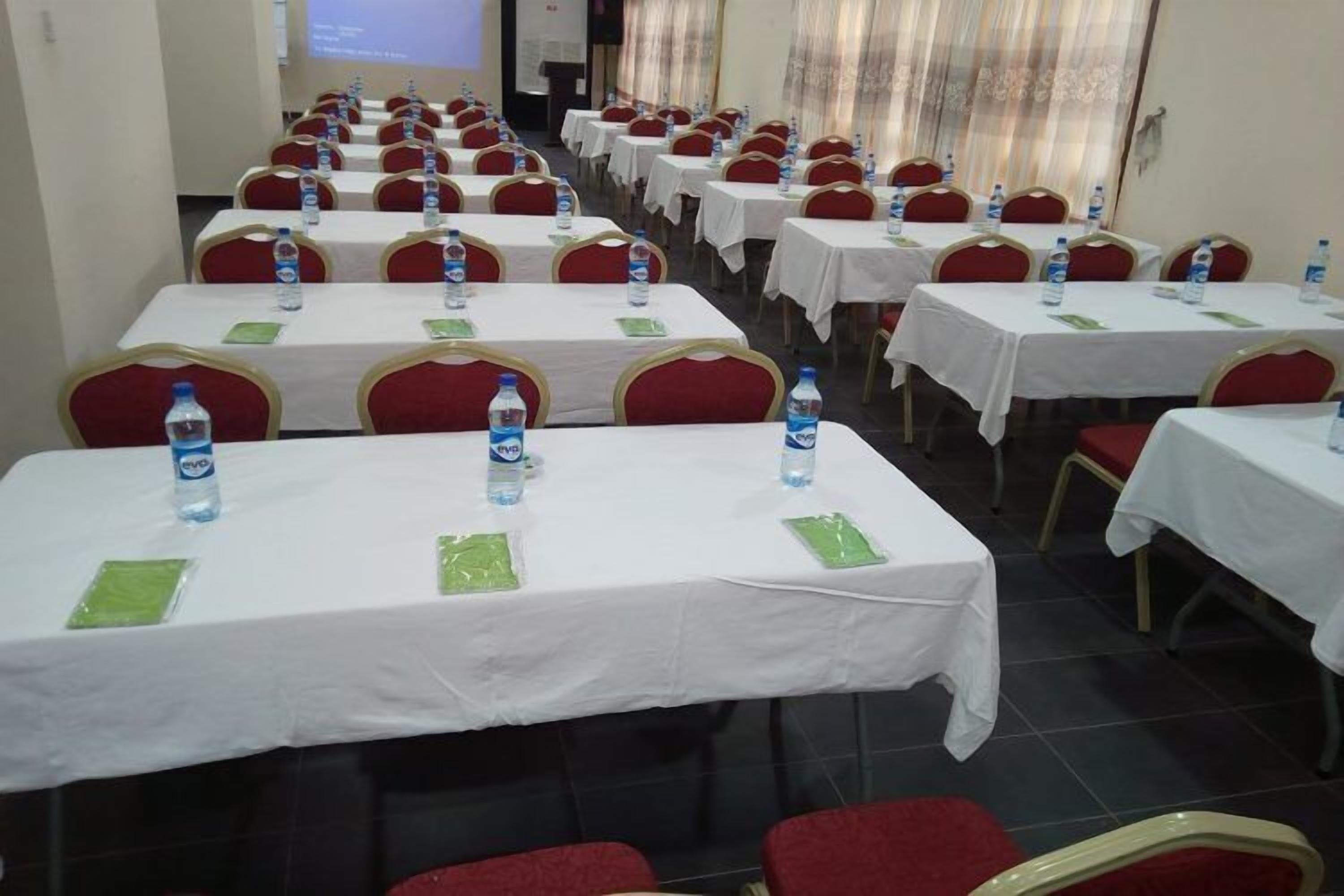 Une salle de conférence avec des rangées de tables, chacune recouverte d’une nappe blanche, et des chaises rouges, ainsi que des bouteilles d’eau.