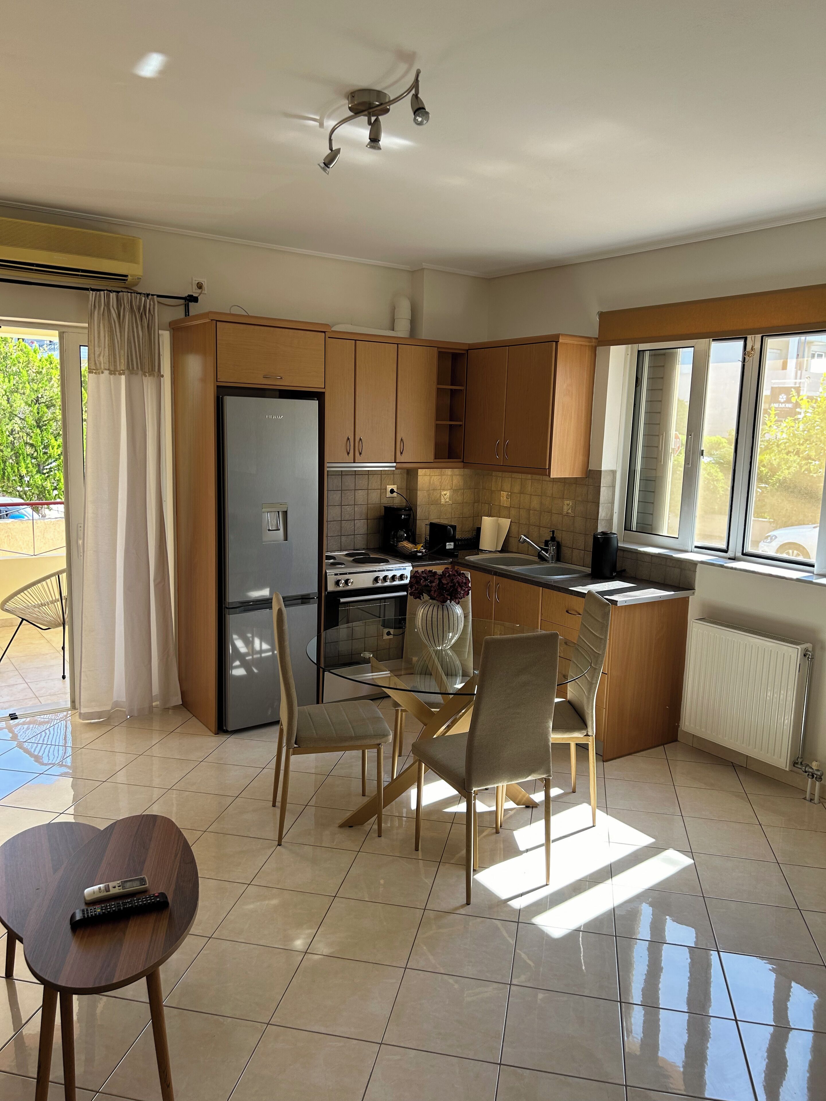 A modern kitchen with wooden cabinets, a built-in oven, and a refrigerator. There is a dining table with chairs and a remote control on a small table.