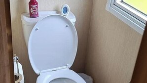A small, compact bathroom with a white toilet, a small window, and a red can on the toilet tank. - Seaside Caravan Retreat in Craig Tara (Ayr)