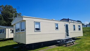 A mobile home with a front porch, a small attached garage, and a windowed side section. - Seaside Caravan Retreat in Craig Tara (Ayr)