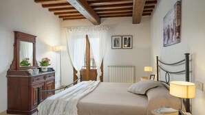 Ein Schlafzimmer mit Holzboden, einem Bett mit weißer Bettdecke, einem Nachttisch mit Lampe, einer Kommode und einem Fenster mit Vorhängen.