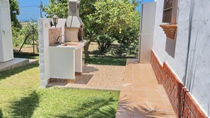 A patio area with a stone-tiled floor, a white outdoor kitchen with a chimney, and a view of a tree-lined landscape.