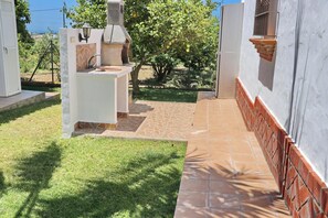 A patio area with a stone-tiled floor, a white outdoor kitchen with a chimney, and a view of a tree-lined landscape.