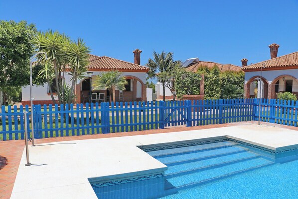 A swimming pool with a blue tiled border, a blue fence, and a building with red-tiled roofs in the background.