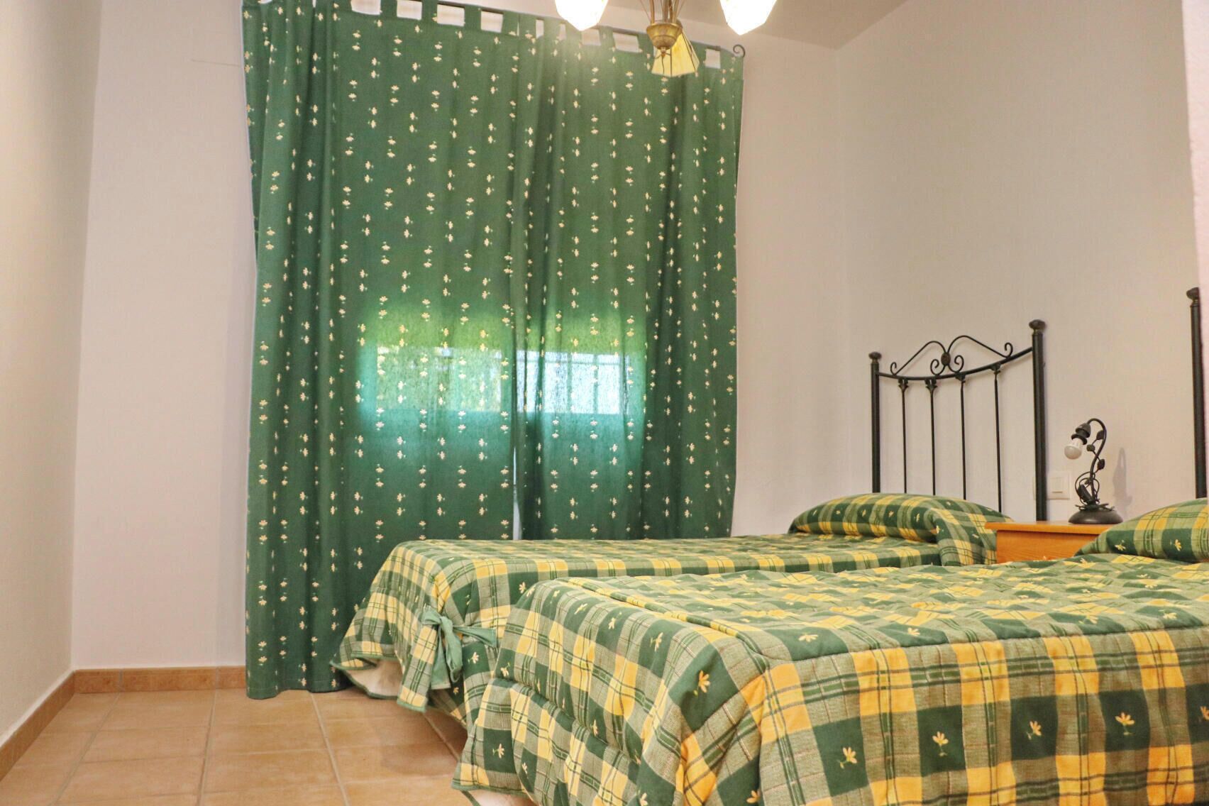 A bedroom with two beds, green curtains with a floral pattern, a metal headboard, and a bedside lamp.