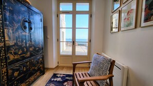 A room with a large cabinet, a patterned chair, a rug, and a door leading to a balcony with a view of the sea. - Magnifique vue Pleine mer à la Perle Marine - La Terrasse (Dinard)