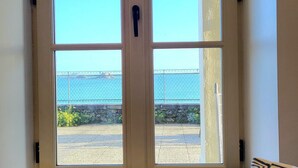A room with a large window overlooking a body of water, a stone wall, and a view of a bridge. There are two pillows on a bench in front of the window. - Magnifique vue Pleine mer à la Perle Marine - La Terrasse (Dinard)