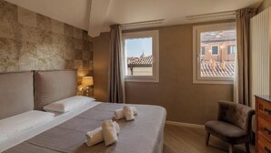 Ein Hotelzimmer mit einem großen Bett, einem Sessel, einem Fenster mit Blick auf Gebäude und einer Lampe auf dem Nachttisch.