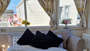 A bedroom with a checkered bedspread, a window with floral curtains, and a view of a building outside. - D&ashortbreaks-stmargaretsbay-parking (Dover)