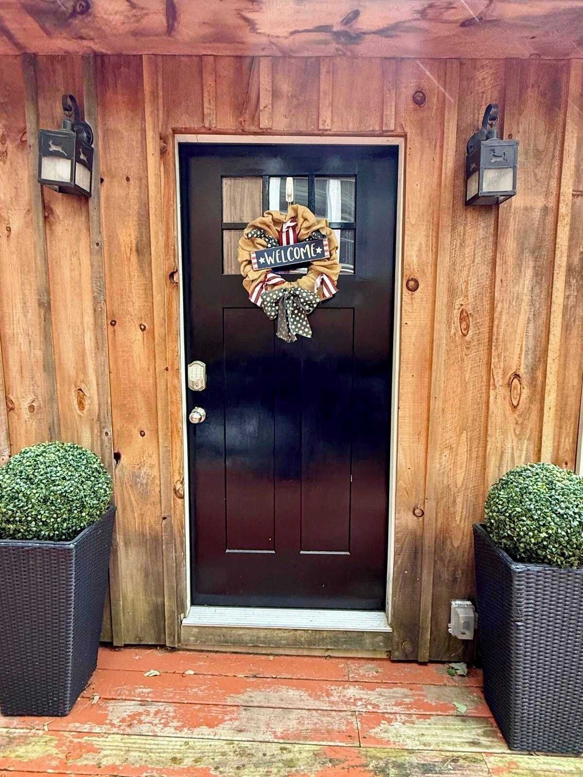 A wooden door with a welcome wreath, two potted plants, and a dark-colored door.