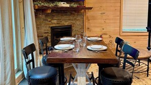 A dining area with a wooden table set for four, a television mounted on a stone wall, and a fireplace below. - Treehouse Duo Lakefront  2 Private Hot Tubs (Belgrade)