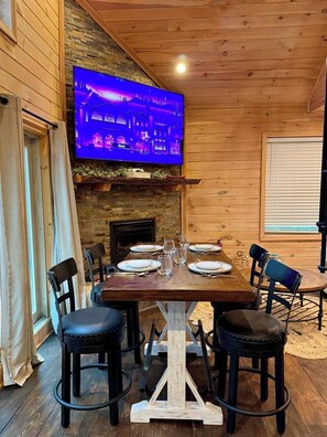 A dining area with a wooden table set for four, a television mounted on a stone wall, and a fireplace below. - Treehouse Duo Lakefront  2 Private Hot Tubs (Belgrade)