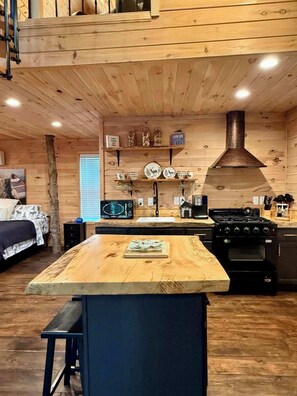 A cabin interior with a kitchen area featuring a wooden island, a stove, and a hanging shelf with decorative items. - Treehouse Duo Lakefront  2 Private Hot Tubs (Belgrade)