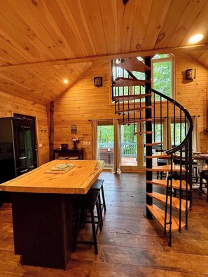A wooden cabin interior with a central island, a spiral staircase, and a large window. - Treehouse Duo Lakefront  2 Private Hot Tubs (Belgrade)