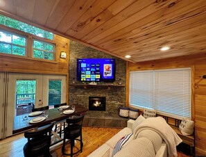 L’intérieur chaleureux d’un chalet comprend un coin repas, une cheminée, un téléviseur fixé au mur en pierre et une fenêtre avec des stores.