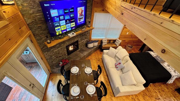 A cozy cabin interior with a stone fireplace, a flat-screen TV mounted on a wall, a dining area with a table set for four, and a comfortable sofa. - Treehouse Duo Lakefront  2 Private Hot Tubs (Belgrade)