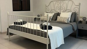 Un lit simple avec un cadre en métal, une table de chevet, une lampe de chevet, une fenêtre et un luminaire fixé au mur.