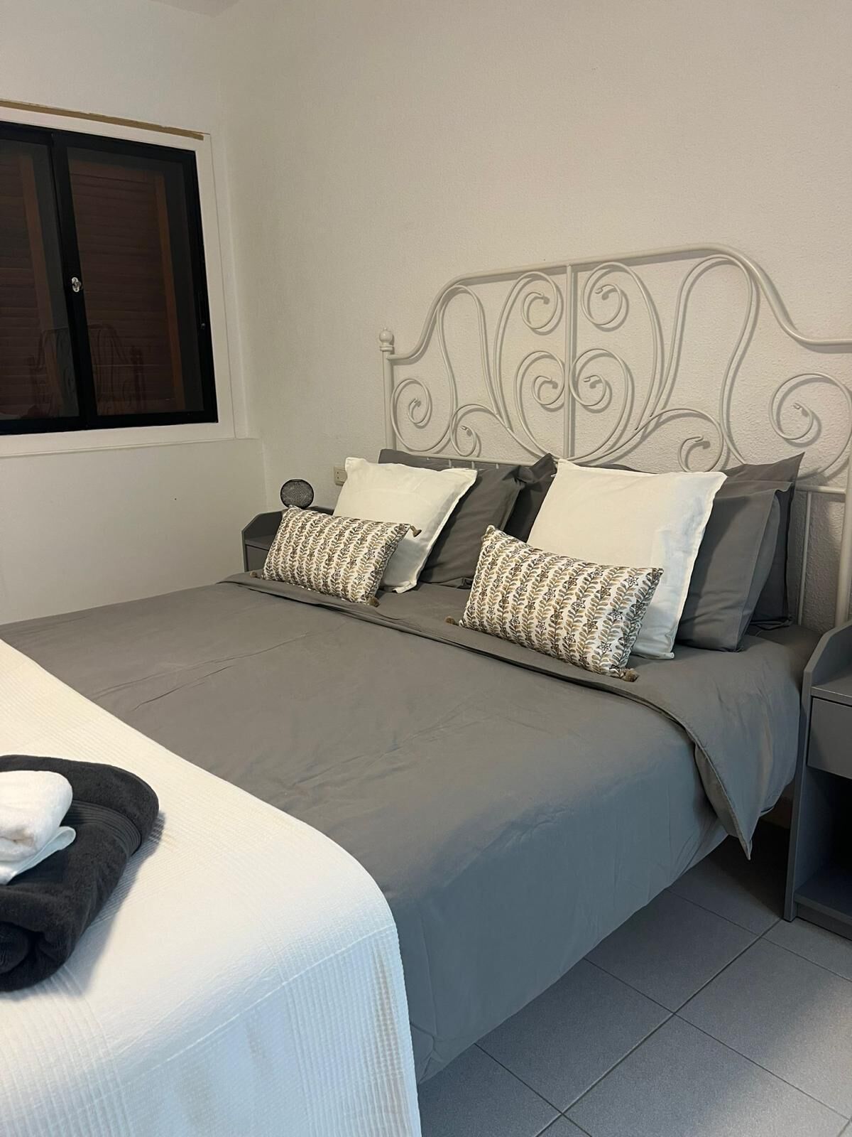 A neatly made bed with a grey bedspread and patterned pillows, a white headboard, and a window with a closed dark blind.