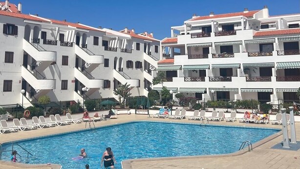 Une piscine entourée de bâtiments blancs aux toits et balcons rouges, avec un ciel bleu dégagé au-dessus.