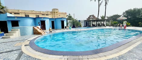 A swimming pool with a tiled edge and a patterned floor, surrounded by a building with blue walls and a covered seating area.