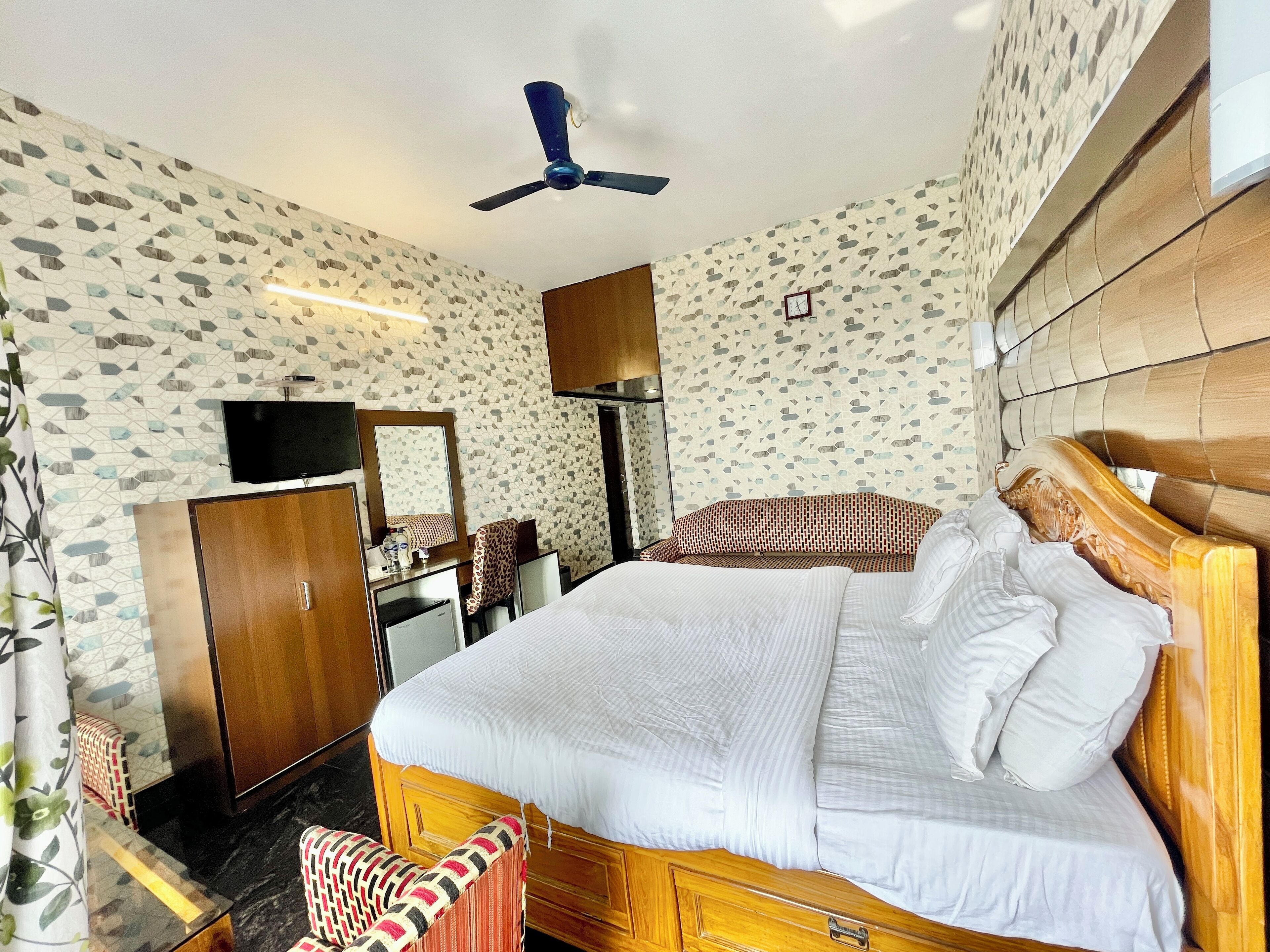 A hotel room with a bed, a desk with a mirror, a television, a wardrobe, and a ceiling fan.