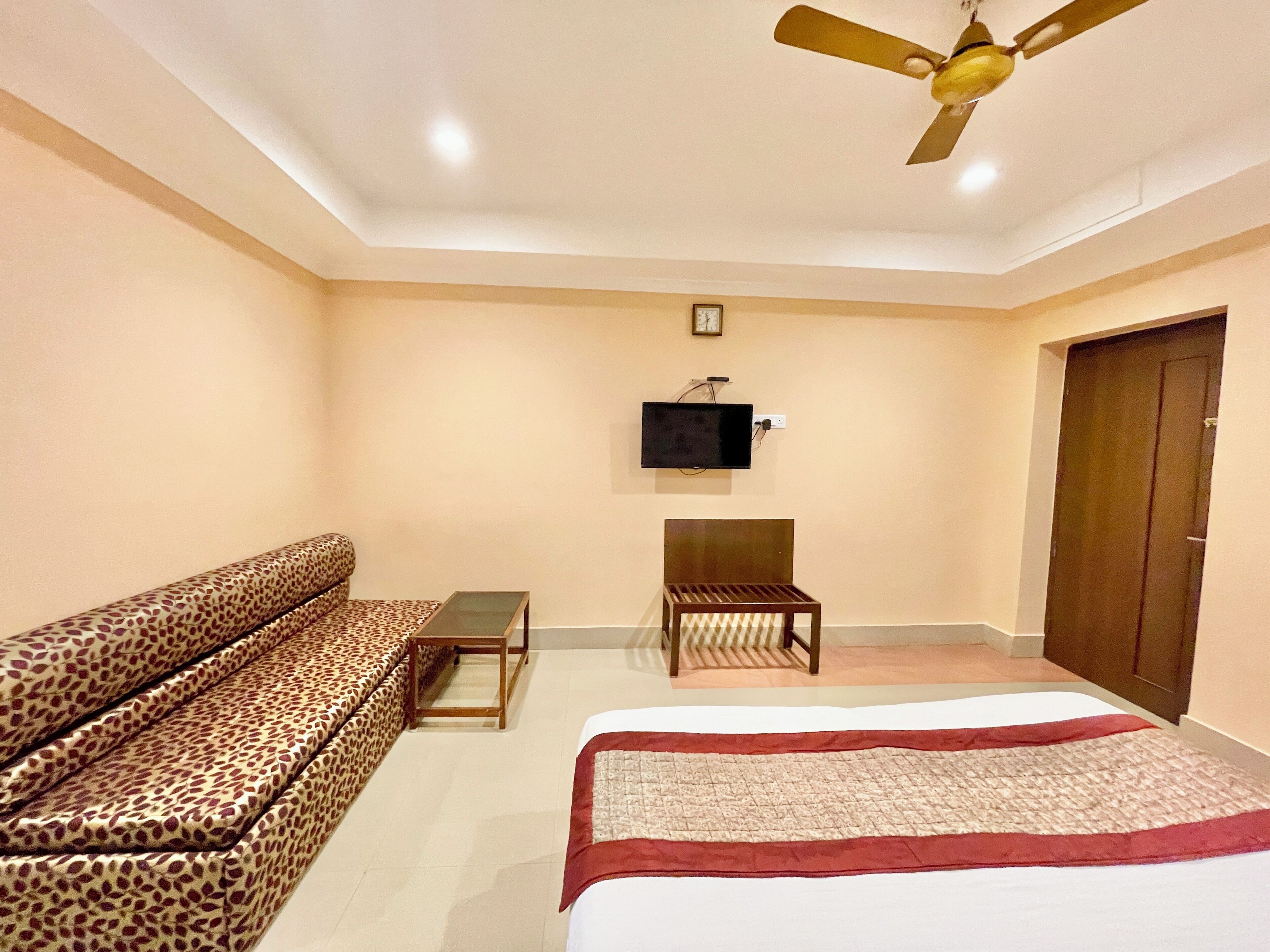 A hotel room with a sofa, a bed, a small table, a chair, a ceiling fan, and a TV mounted on the wall.