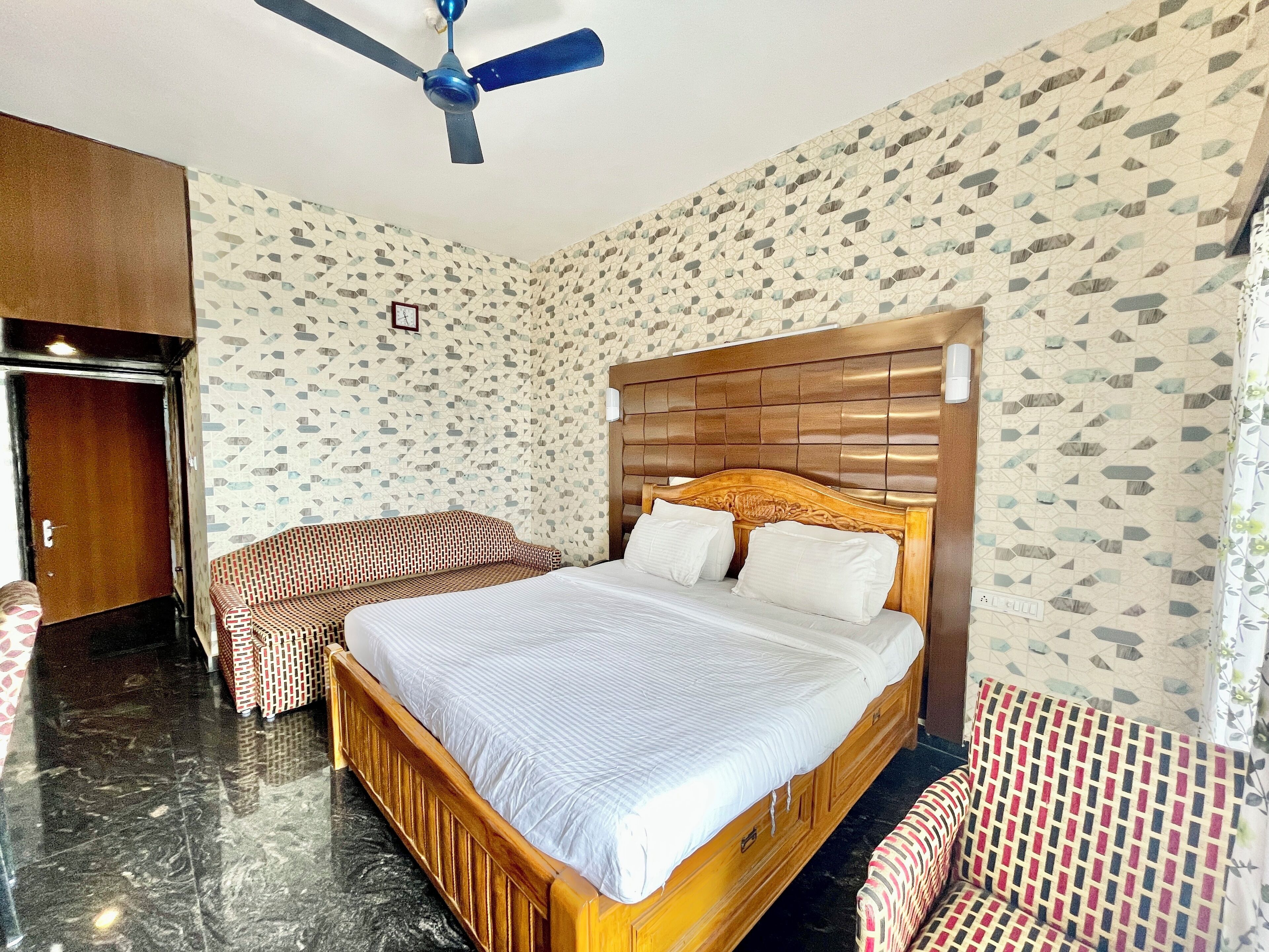 Quarto de hotel com cama grande, cabeceira de madeira, ventoinha no teto e paredes com padrão.