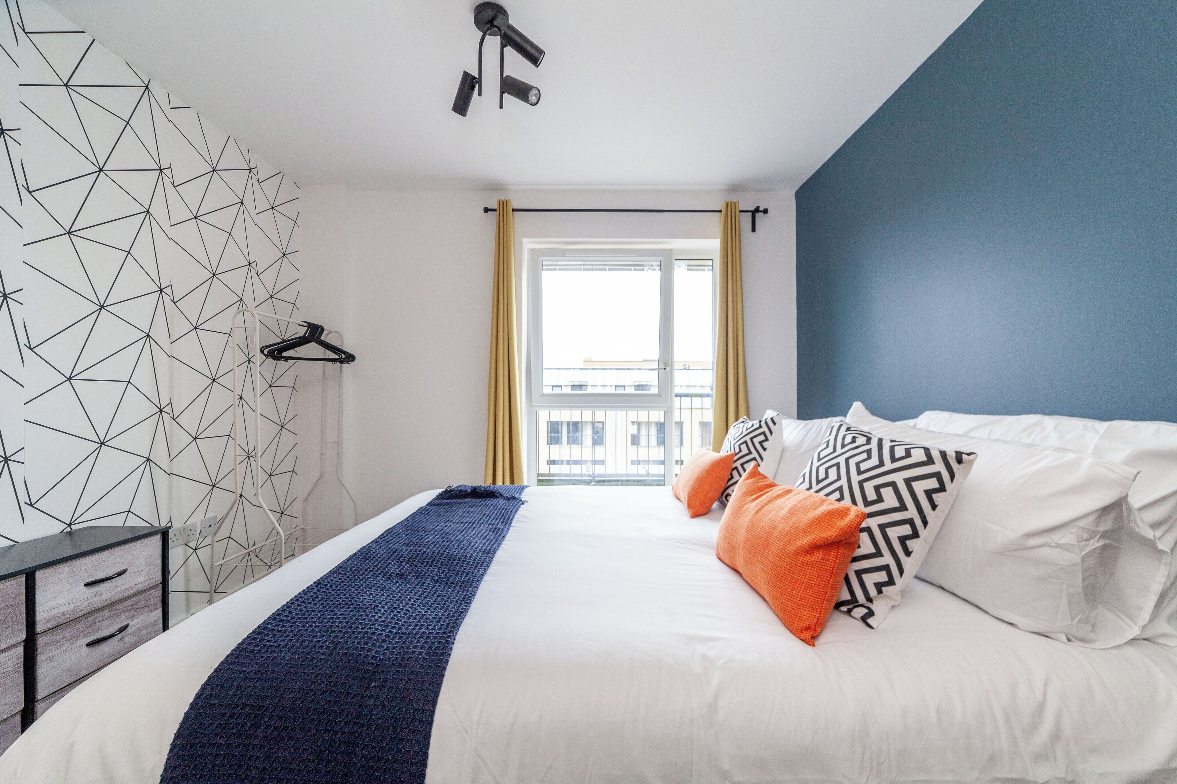 A modern hotel room with a geometric-patterned wall, a bed with white and patterned pillows, a blue blanket, and a view of a building through a window with yellow curtains.