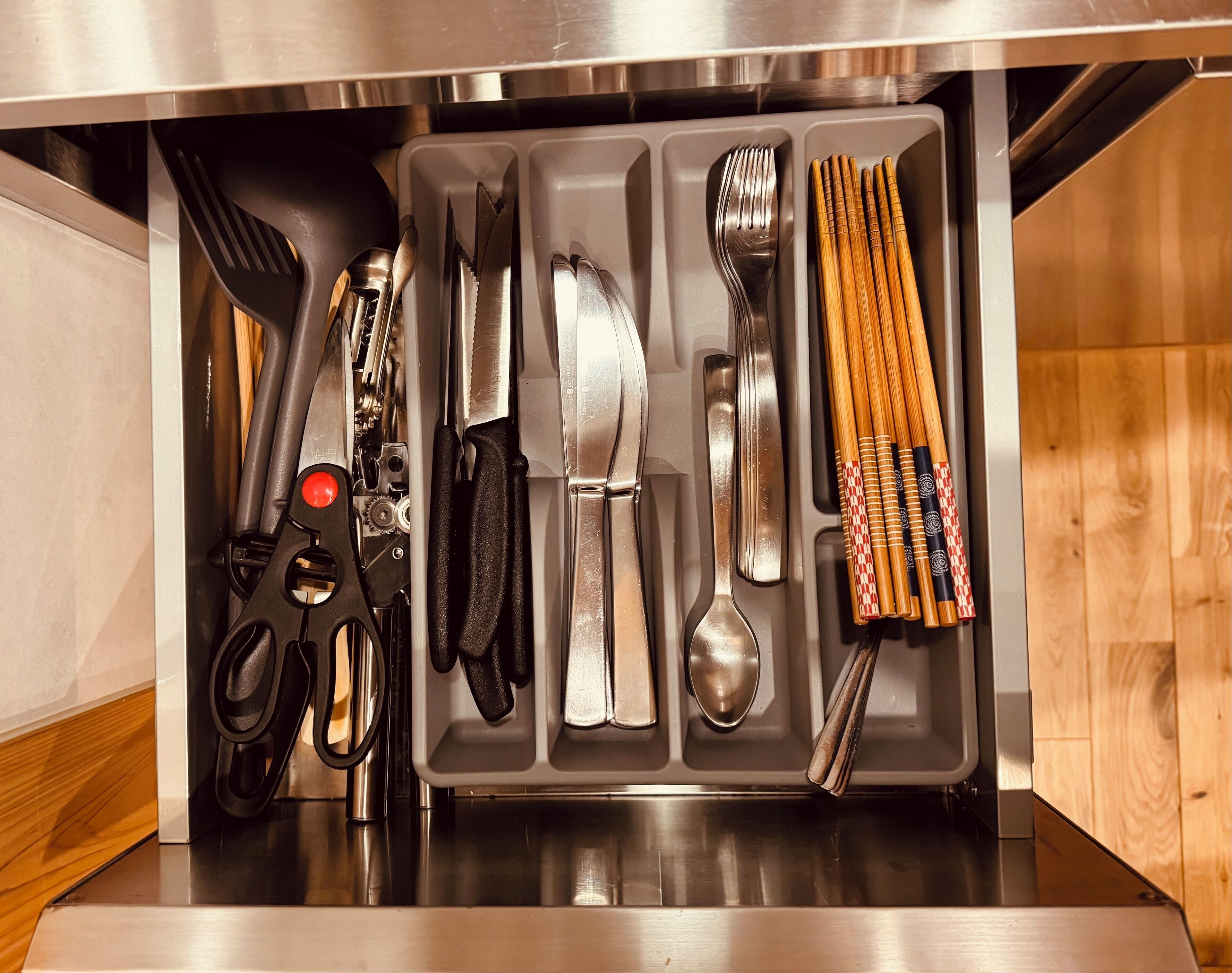 A kitchen drawer with utensils and cutlery including a spatula, tongs, knives, a spoon, and chopsticks.