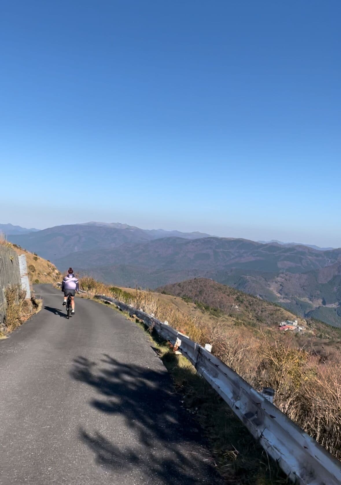 A cyclist on a paved road with a clear view of distant mountains and a small building in the valley.