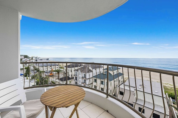 A balcony with a wooden table and white chairs, overlooking a beach and residential buildings.