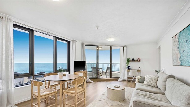 A spacious living room with a large window offering a sea view, a balcony with a table and chairs, and a cozy seating area with a sofa and a round coffee table.