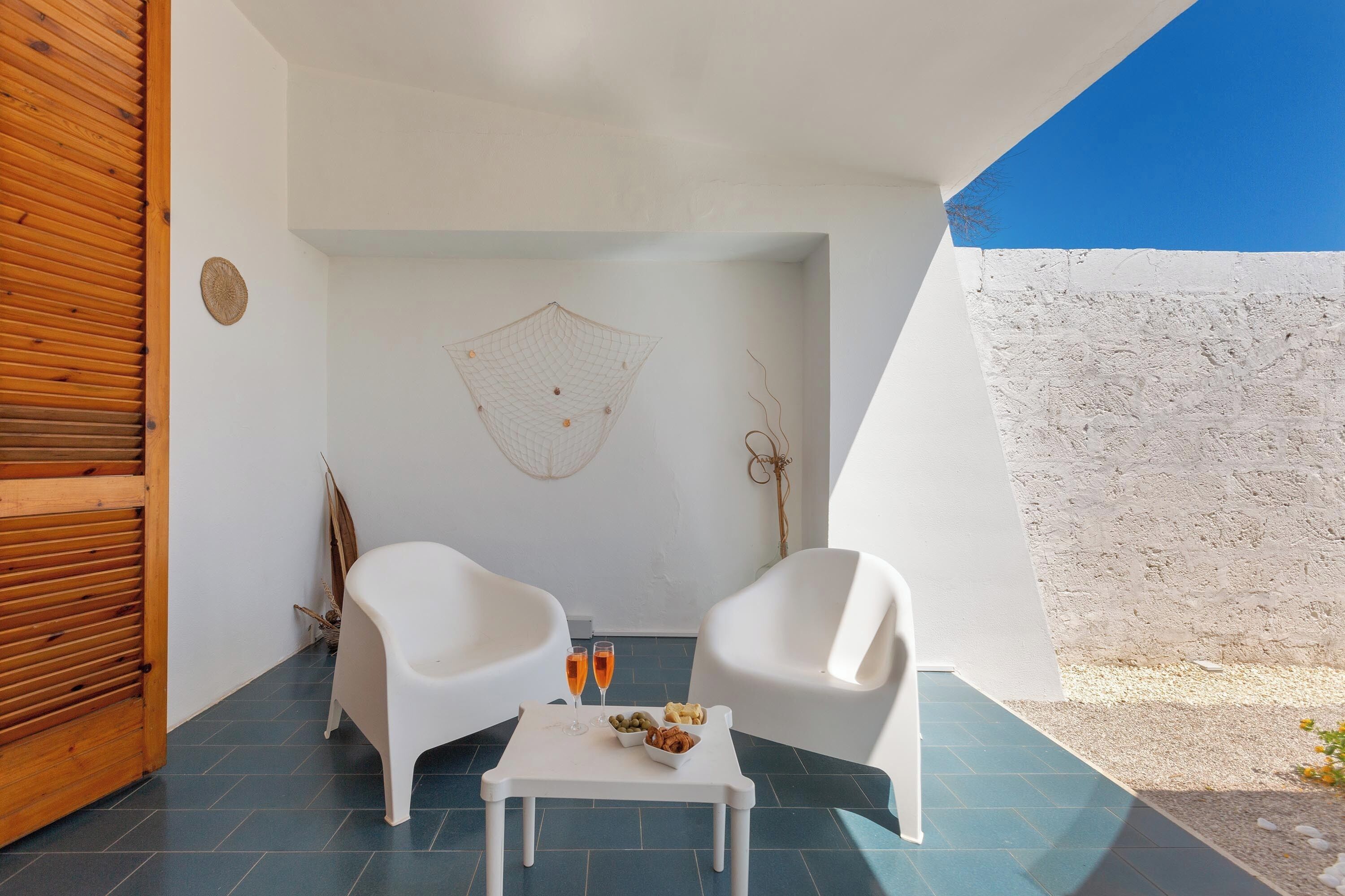 A small, private outdoor seating area with two white chairs and a table, featuring a blue tiled floor and a white wall with a decorative net.